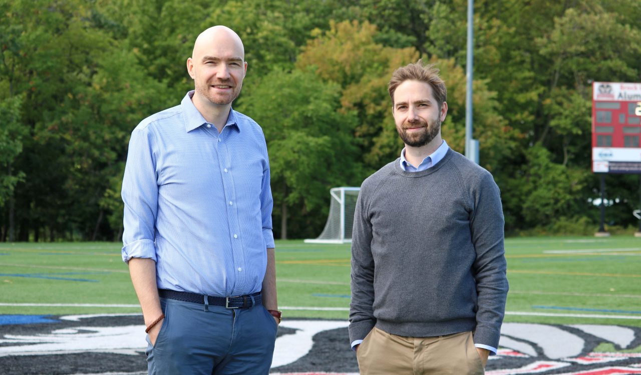 Two men in business casual attire stand on a sports field with a forested area in the background.