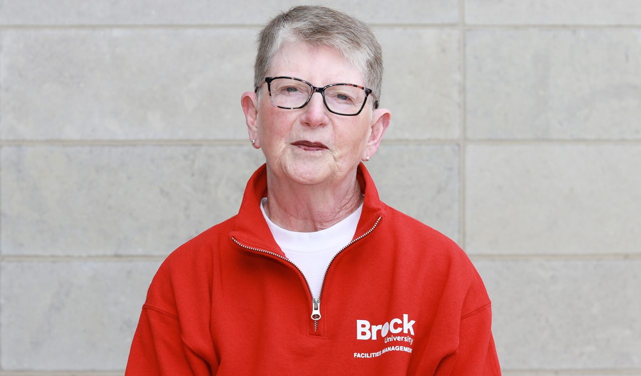 A headshot of a woman standing against a grey stone wall wearing a red zip-up sweater with the words “Brock University Facilities Management” written in white.