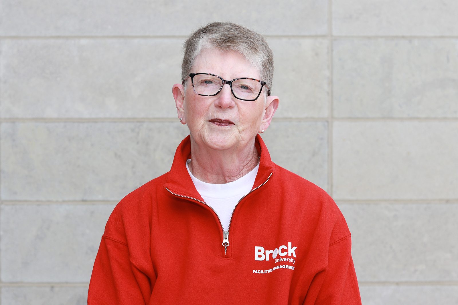 A headshot of a woman standing against a grey stone wall wearing a red zip-up sweater with the words “Brock University Facilities Management” written in white.
