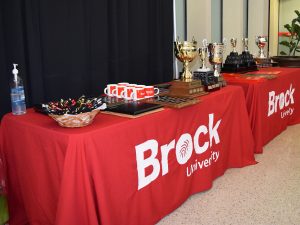 Two tables draped in red tablecloths with “Brock University’ in white display several trophies, plaques, mugs, pens, scrolls and a bottle of hand sanitizer.