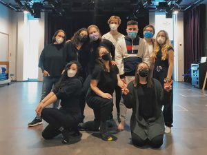 A group of 10 people wearing face masks stand together posing for the camera in an indoor studio space with a black background and theatre equipment lining the walls of the room. Natural light spills in from the windows.