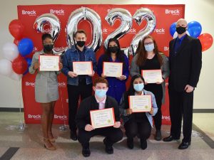 Seven people stand together in two rows in front of a red backdrop with white ‘Brock University’ repeated several times. Red, white and blue balloons flank them on either side. Silver ‘2022’ balloons float behind them. All but one person is holding a paper certificate with their name printed on it. Everyone is wearing a mask.