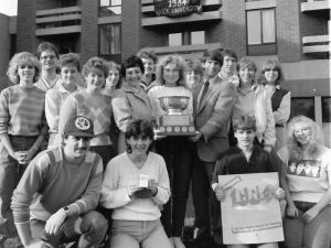 Seventeen people gather outside in front of a university residence building to pose for a photo. Most people are standing and four people are on their knees at the front of the group. Three of the people standing hold a large trophy. One person who is kneeling holds a poster that reads ‘Homecoming 1984.’