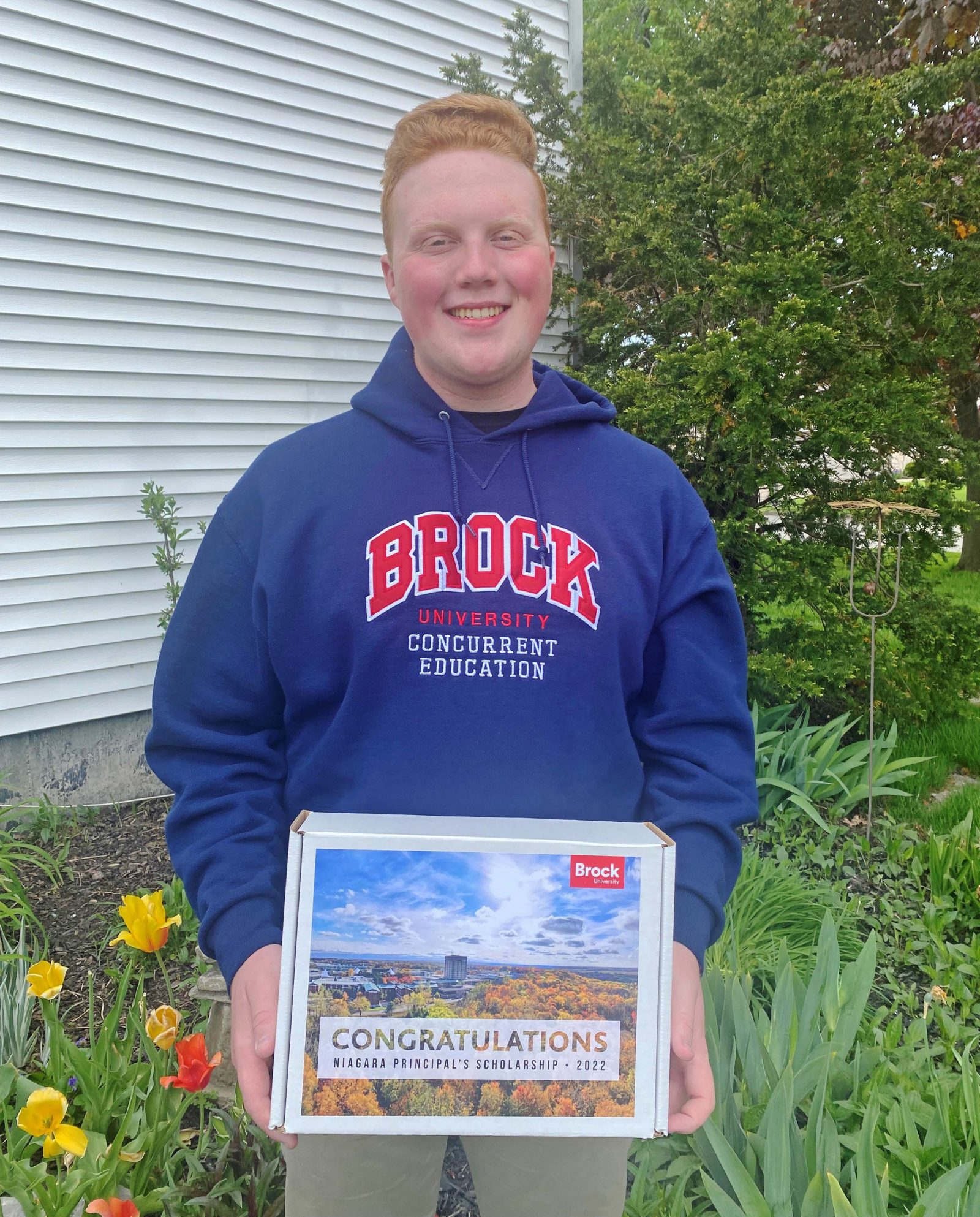 A young man in a blue Brock University hoodie holds a box that says congratulations on it.