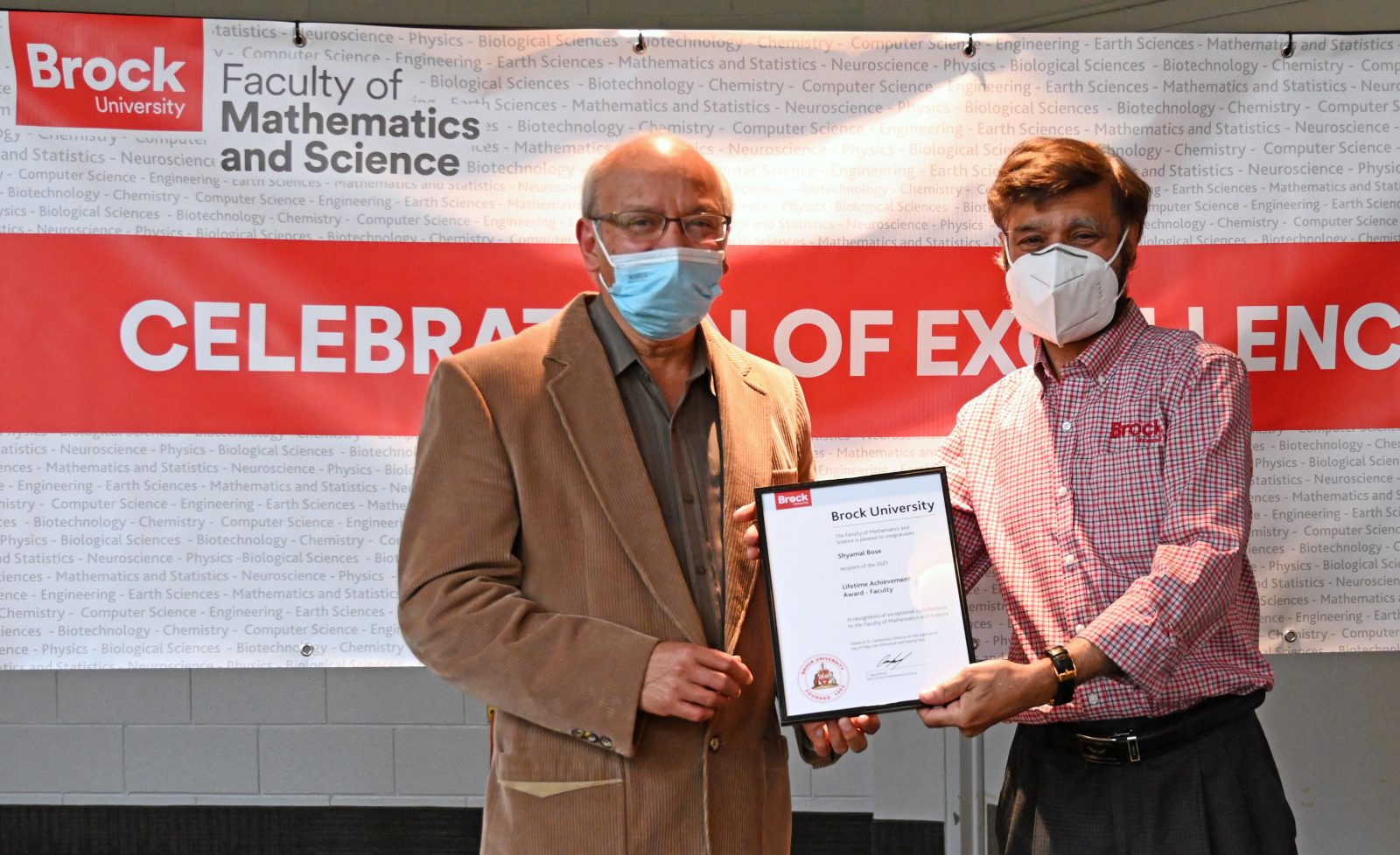 Two men stand in front of event banner jointly holding a framed award certificate and smiling for the camera. Both men wear medical masks and semi-formal attire.