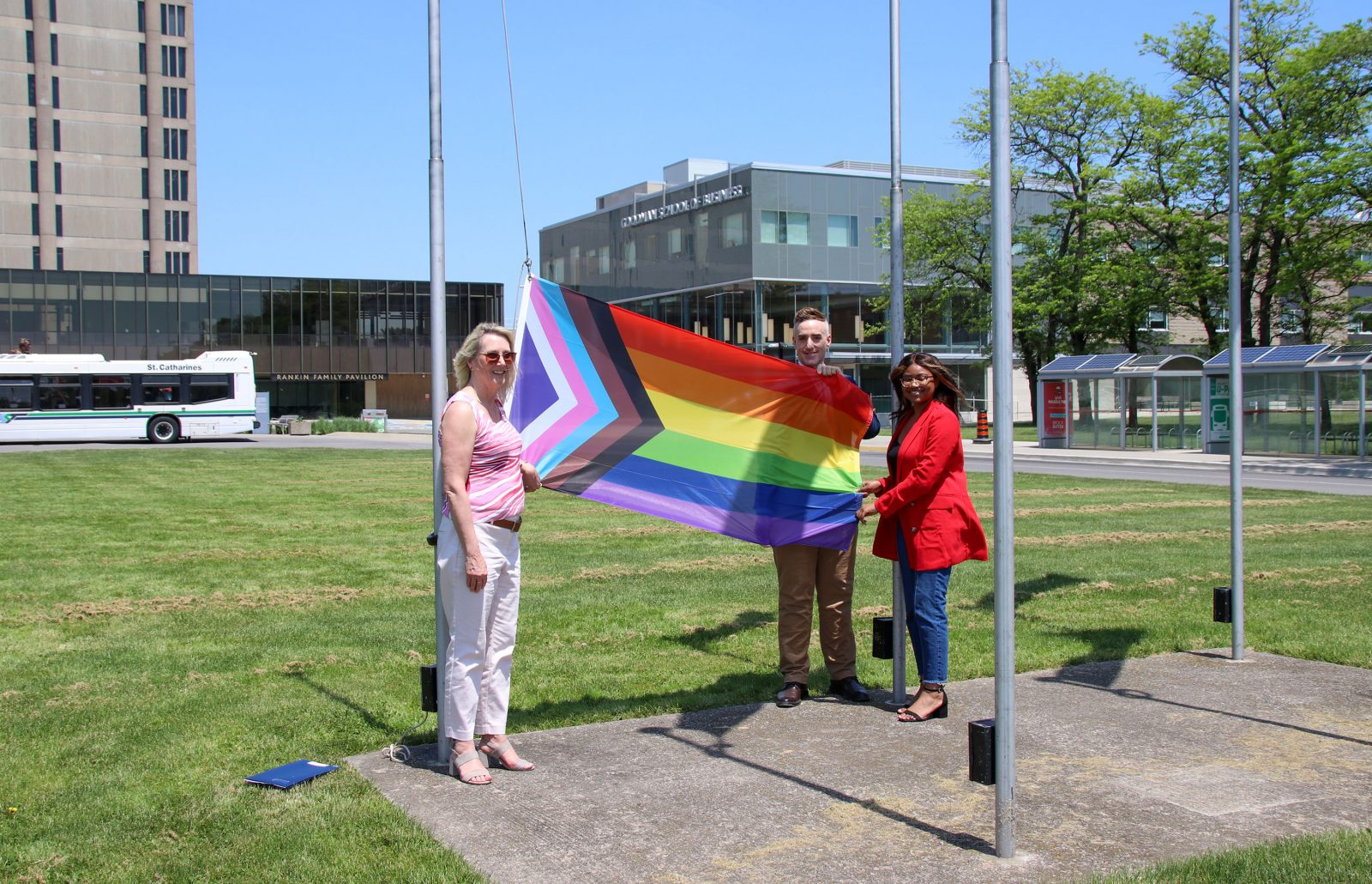 A woman in a pink shirt, a man in brown pants and a woman in a red blazer hold an All-inclusive flag, prior to it being raised.