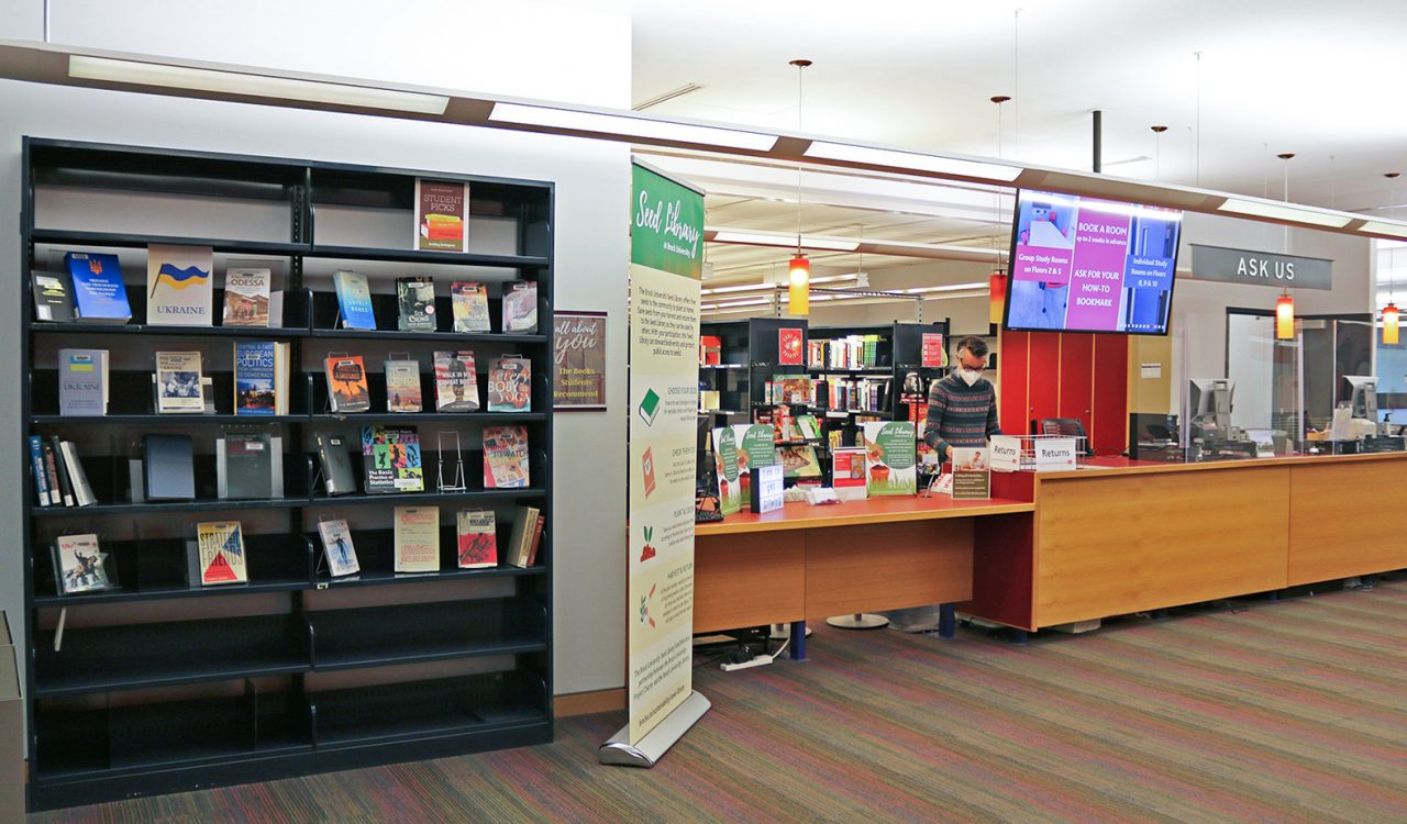 A person stands behind a long counter with their head down, focused on a task. Above the counter is a large screen with information about booking group study rooms. A sign that reads ‘Ask Us’ hangs from the ceiling. To the left of the counter is a large bookshelf with two dozen books on display and a tall pullup banner with information about a Seed Library.