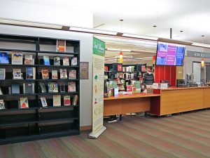 A person stands behind a long counter with their head down, focused on a task. Above the counter is a large screen with information about booking group study rooms. A sign that reads ‘Ask Us’ hangs from the ceiling. To the left of the counter is a large bookshelf with two dozen books on display and a tall pullup banner with information about a Seed Library.
