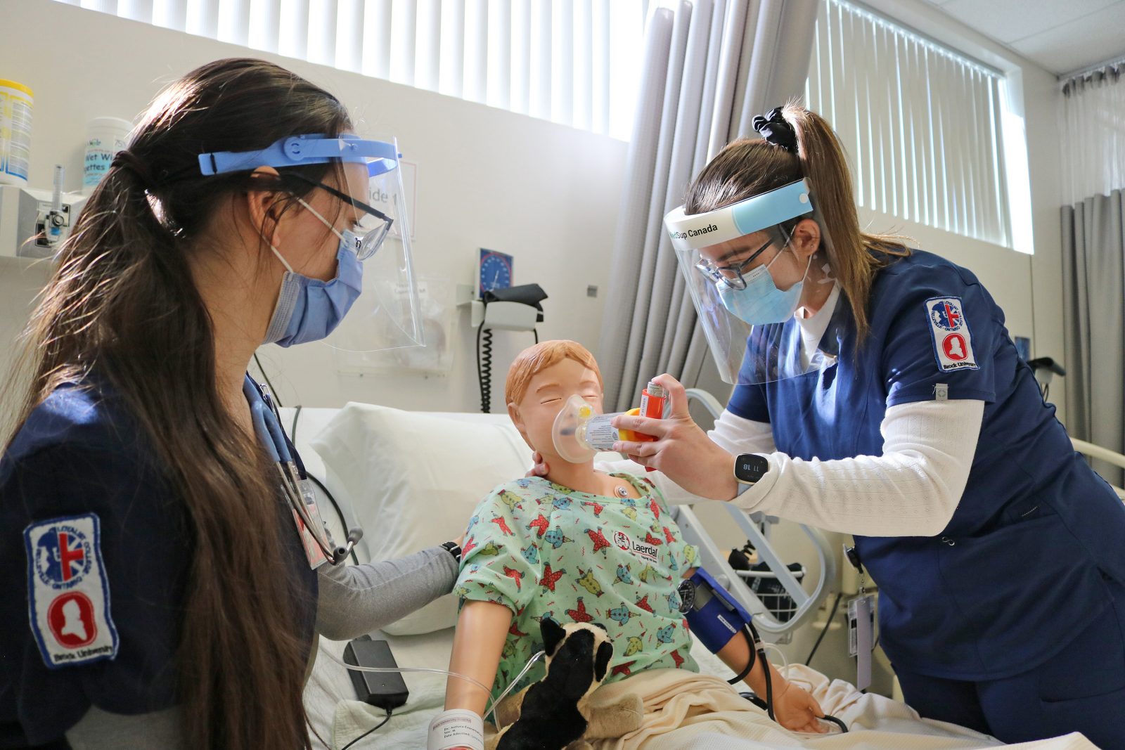 Two nursing students wearing medical masks and face shields use a breathing device on a simulator that looks like a human patient in one of Brock's Nursing labs.