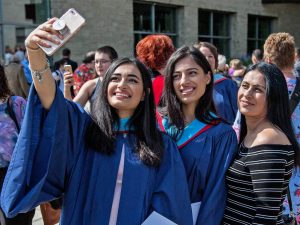 Two graduates from Brock University's Faculty of Education take a selfie outside with a family member during Brock University’s 2019 Spring Convocation. The graduates are wearing blue gowns with blue ceremonial hoods around their necks and shoulders. One of the graduates holds a phone high in the air to snap the photo. Behind the group of three are several other graduates and their family members.