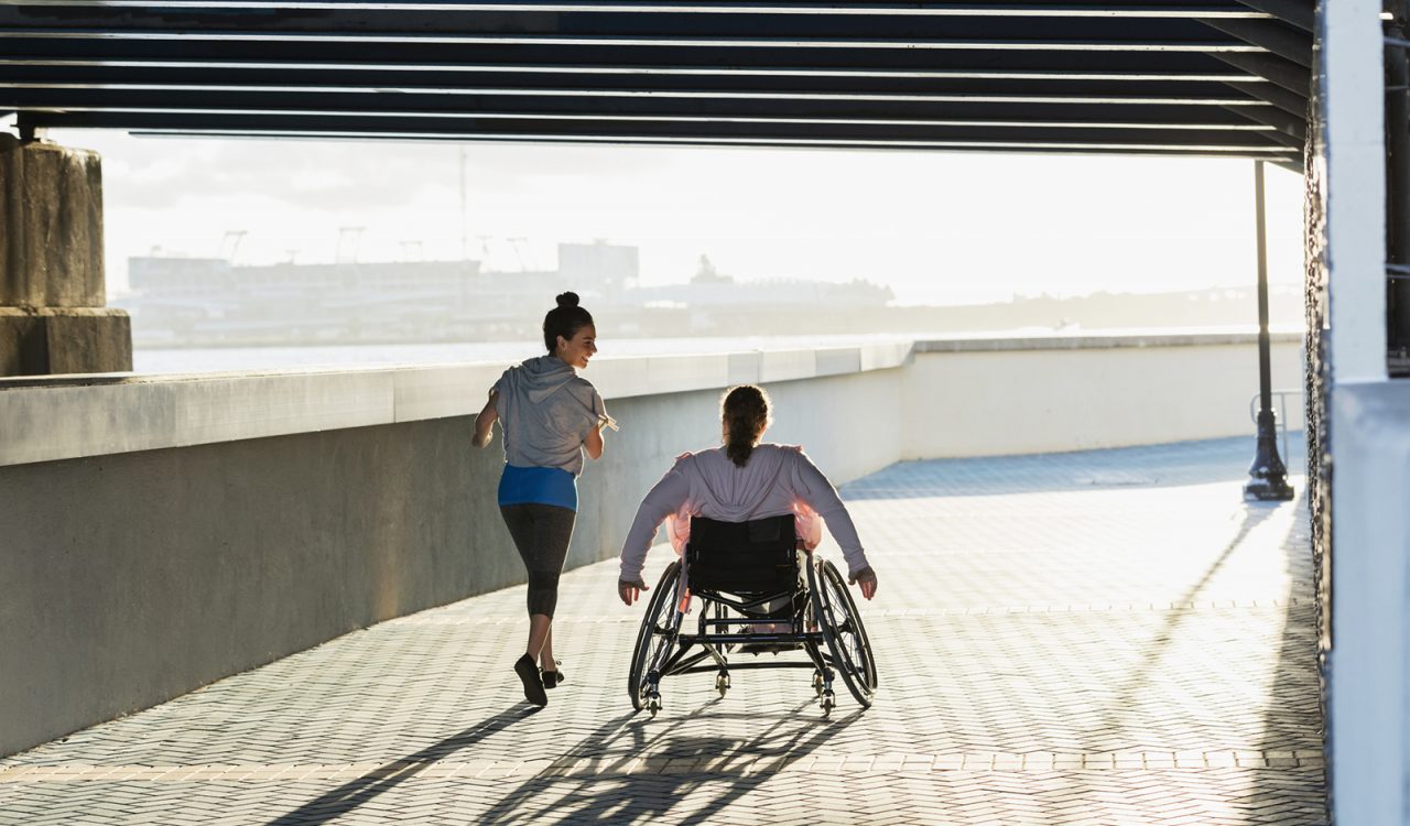 A rear view of two people exercising along a city waterfront. One person is jogging and speaking to her friend, who is rolling beside her in a wheelchair