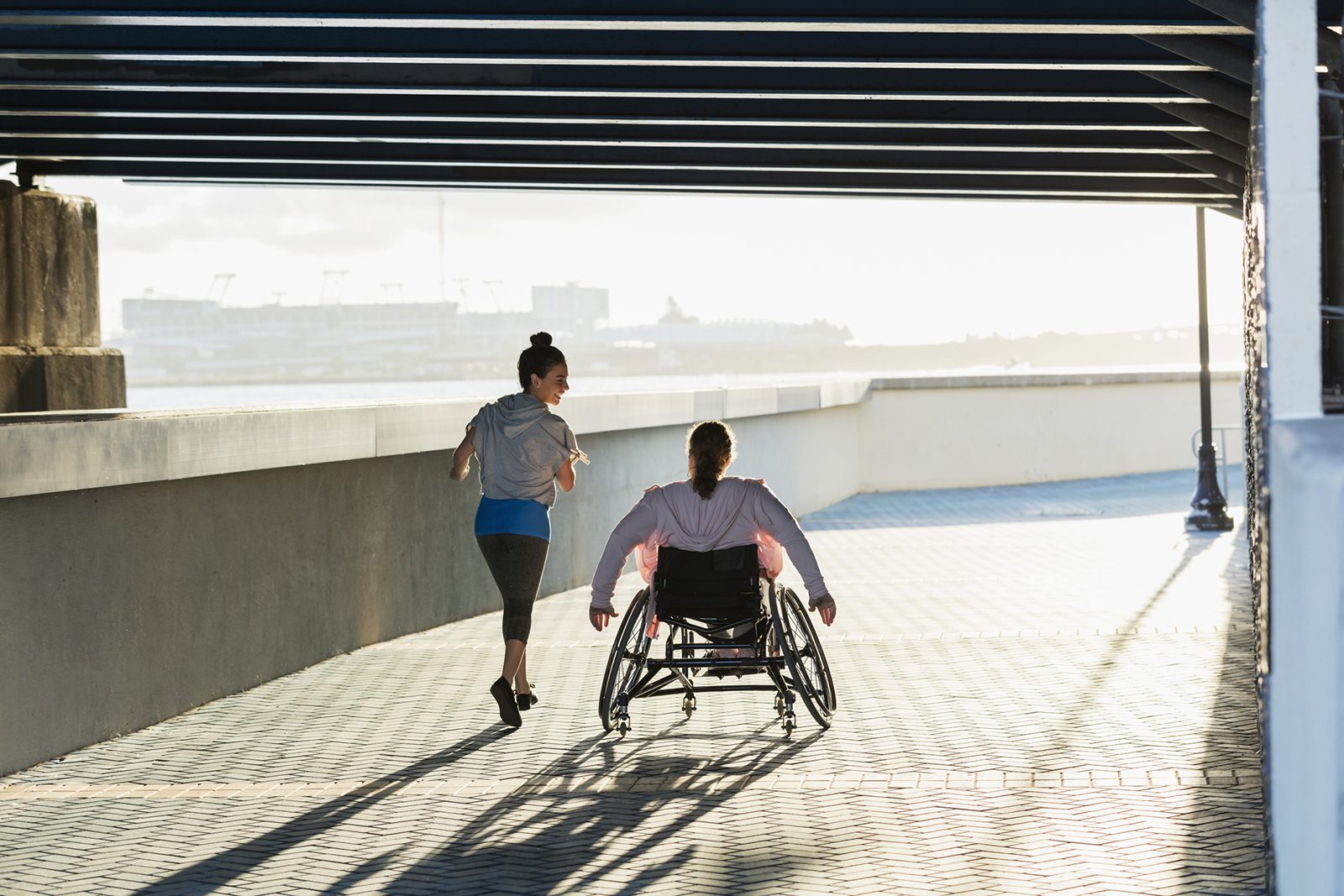 A rear view of two people exercising along a city waterfront. One person is jogging and speaking to her friend, who is rolling beside her in a wheelchair