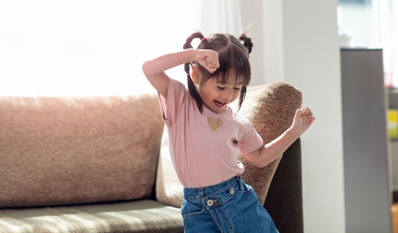A child in a pink T-shirt with pigtails smiles and dances in front of a couch in a sunny room.