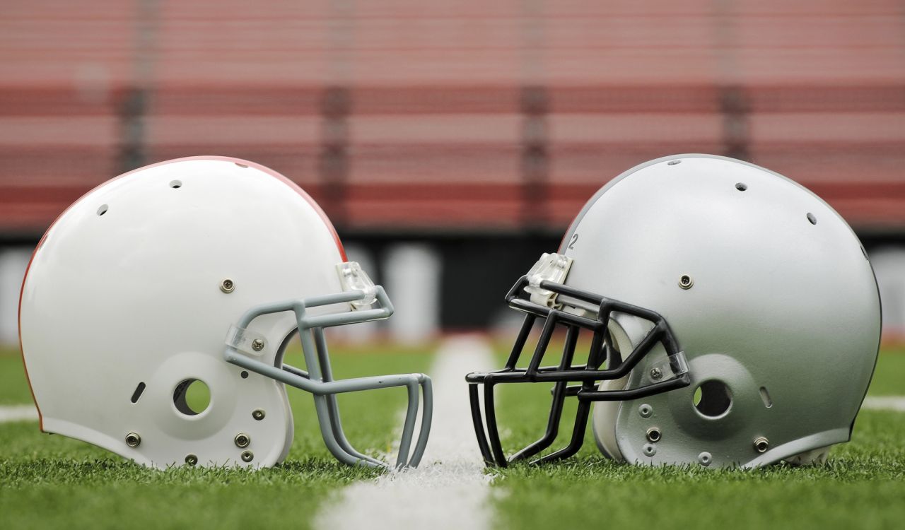An empty white football helmet and grey football helmet face each other on the green grass of a football field.