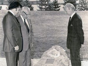 Three men in suits stand around a large rock with a plaque adhered to it. The rock sits on gravel stone. In the background is a small grass hill and several needled trees.
