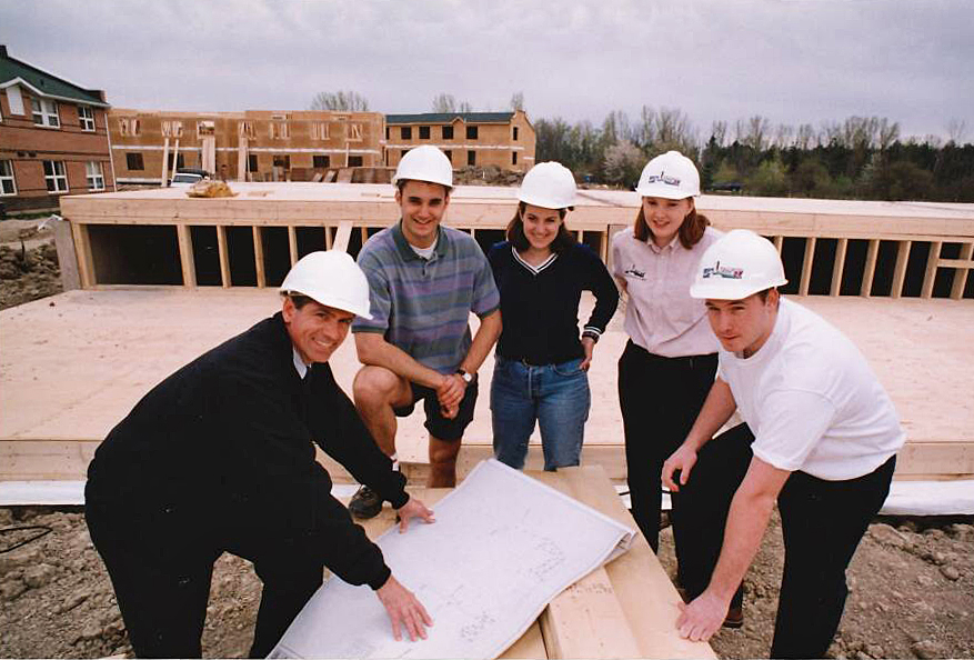 A group of five people wearing white hard hats form a horseshoe around a wooden table with large blueprints displayed on it.