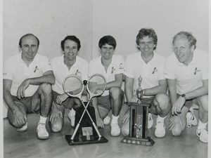 Four squash players kneel on the floor with two trophies in front of them.