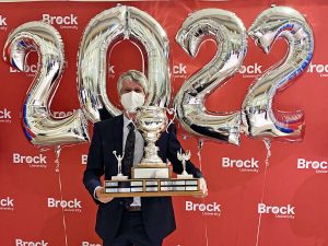 A man holding a large trophy stands in front of a red backdrop with white ‘Brock University’ repeated several times. Silver ‘2022’ balloons float behind him.