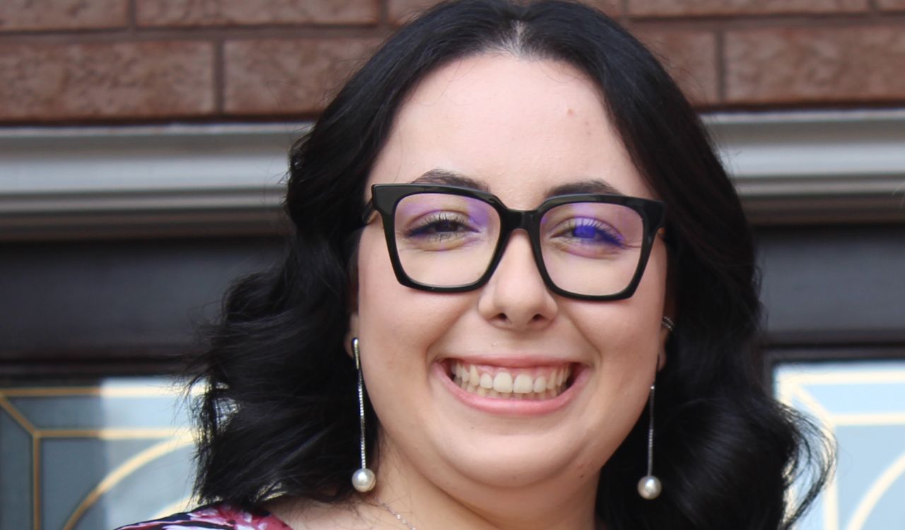 A portrait of Brock student Lucia Marchionda, who has long curled black hair and black thick-framed glasses. She is wearing a navy blue blouse with a pink floral print.