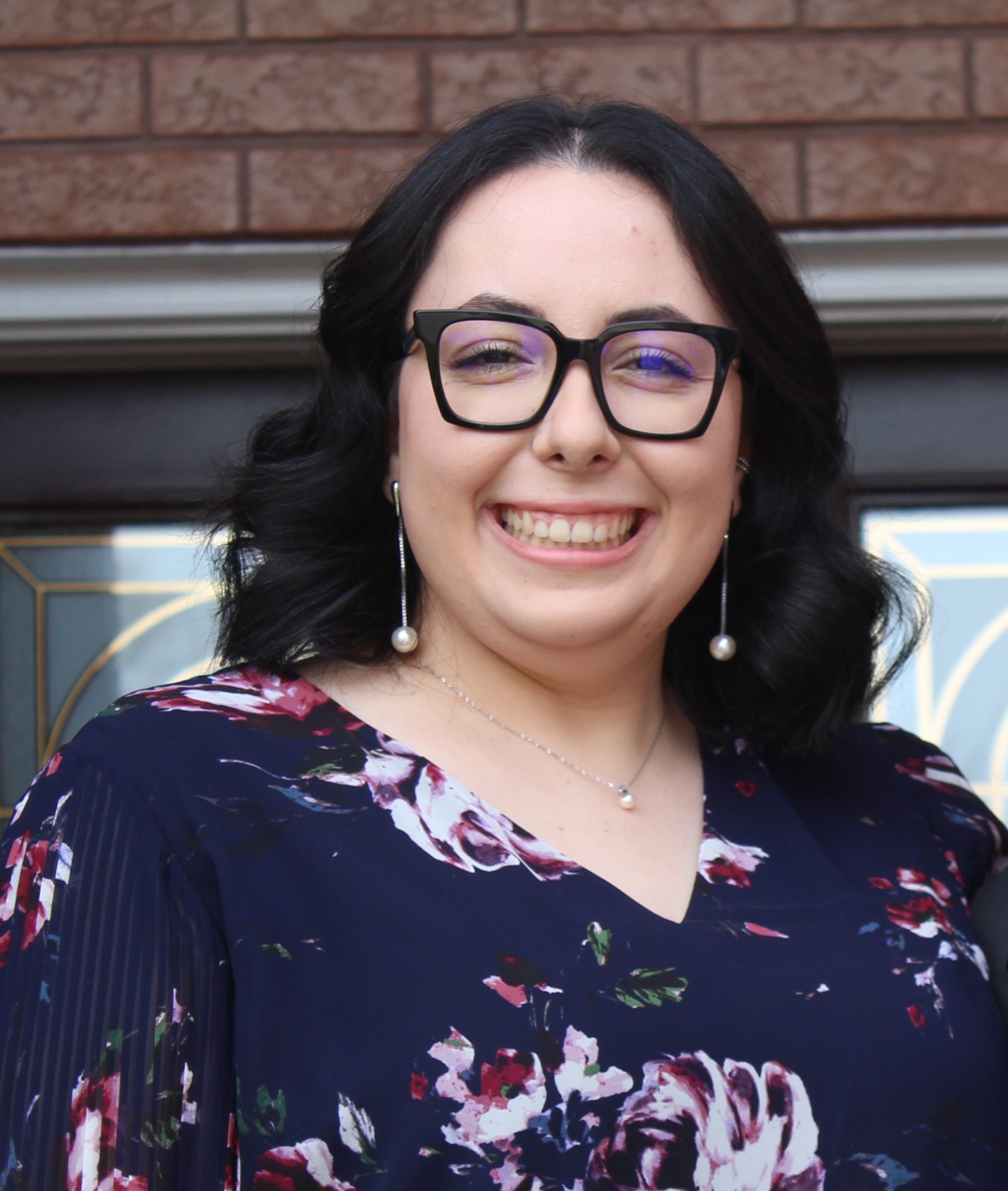 A portrait of Brock student Lucia Marchionda, who has long curled black hair and black thick-framed glasses. She is wearing a navy blue blouse with a pink floral print.