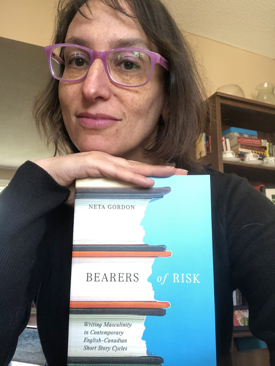 A woman with brown hair and glasses rests her hand on the cover of the book Bearers of Risk.