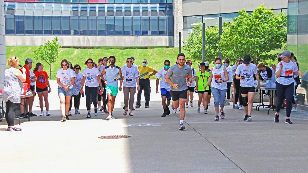 Thirty people line up at a starting line for a community fundraising run. Their arms and legs are in motion as they start the race. In the background is a large grass hill and building.