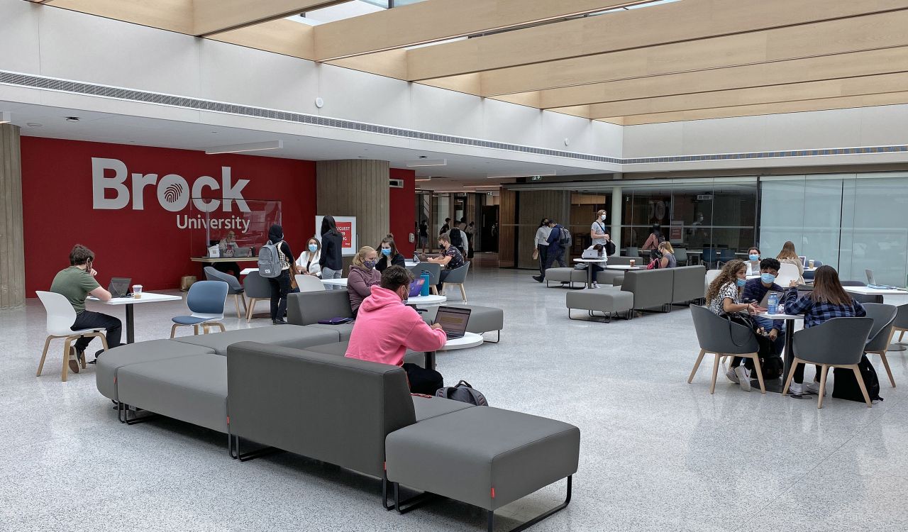 Students site on a series of grey couches underneath a skylight with a large red wall that reads 'Brock University' in white letters in the background.