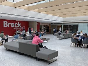 Students site on a series of grey couches underneath a skylight with a large red wall that reads 'Brock University' in white letters in the background.