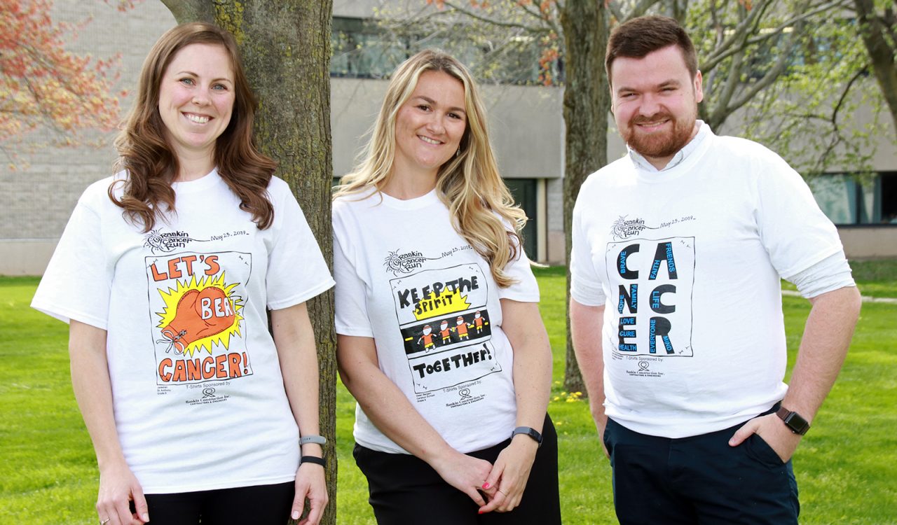 Three people stand side by side outside. One person leans on a tree trunk. They are each wearing white T-shirts with text that reads ‘Rankin Cancer Run.’