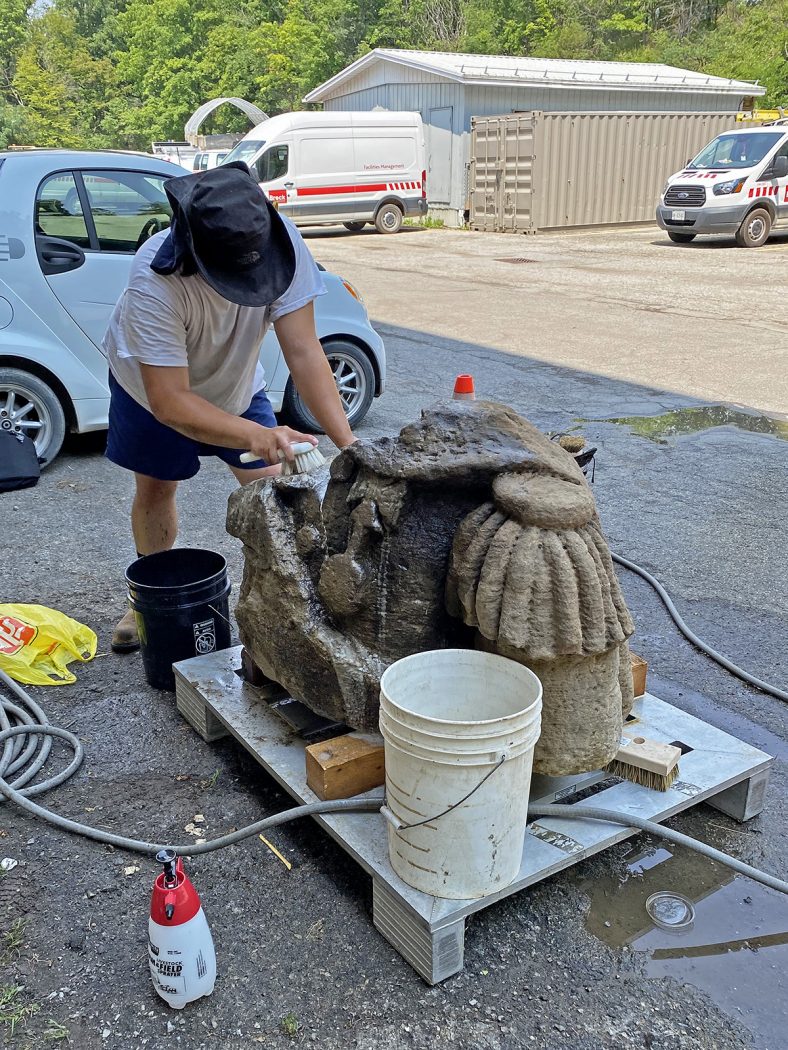 A person wearing a brimmed hat bends over to use a cleaning brush to scrub a stone torso. A long hose is uncoiled on the ground and a large white bucket sits next to the stone. A small container of cleaning chemicals is on the ground.