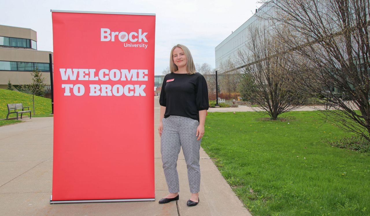 A woman in a black shirt stands next to a large red banner that says, ‘Welcome to Brock.’