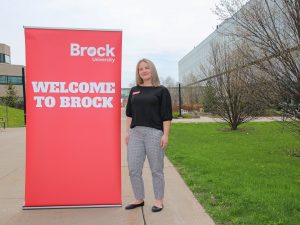 A woman in a black shirt stands next to a large red banner that says, ‘Welcome to Brock.’