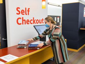 A person picks up a book from several placed on a counter to use at a library self-checkout machine.