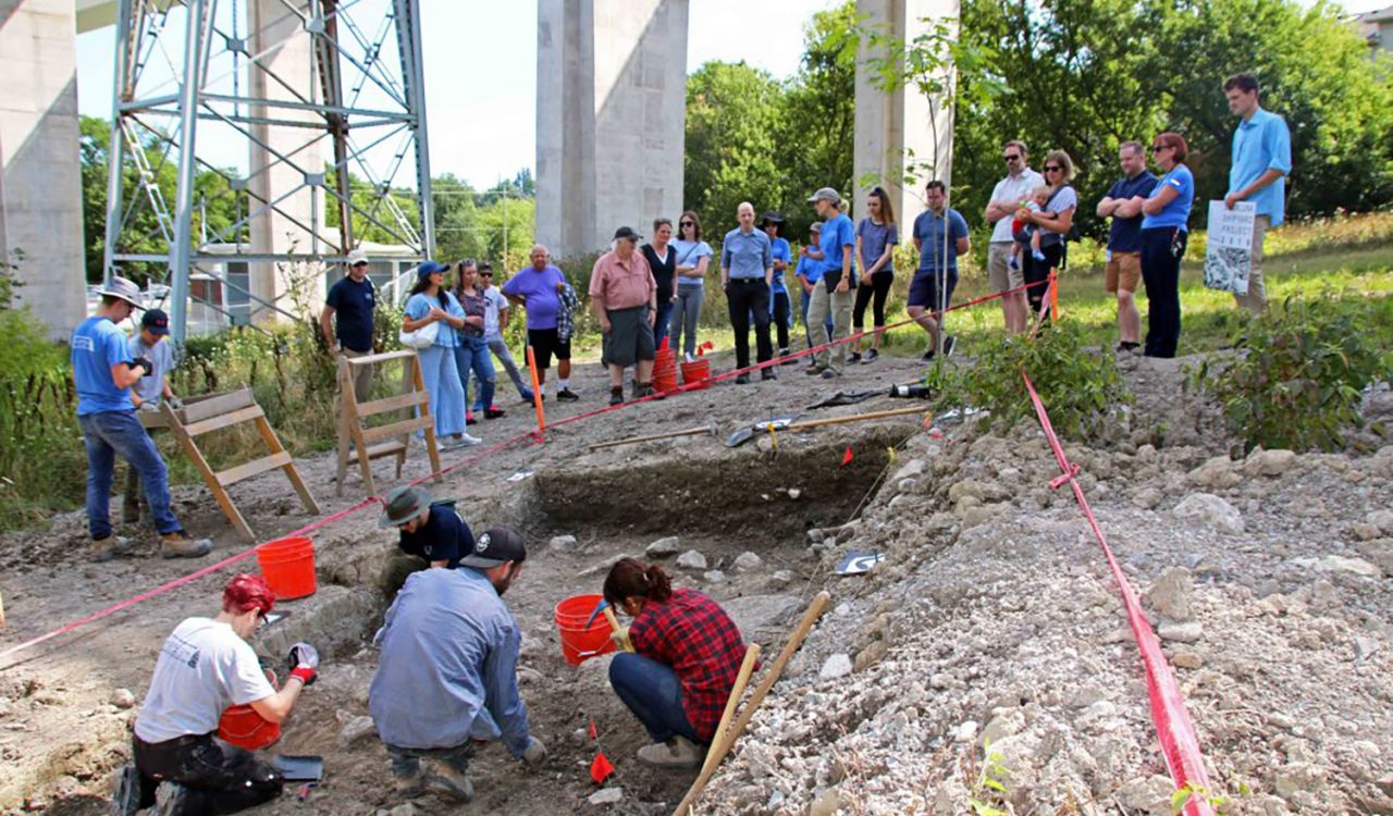 Four students crouch in a shallow, rectangular hole in the ground with buckets and pickaxe. Two students stand at the left of the pit with waist-high screens to sift soil. More than a dozen members of the public watch on from behind a red safety line.