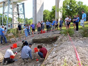 Four students crouch in a shallow, rectangular hole in the ground with buckets and pickaxe. Two students stand at the left of the pit with waist-high screens to sift soil. More than a dozen members of the public watch on from behind a red safety line.