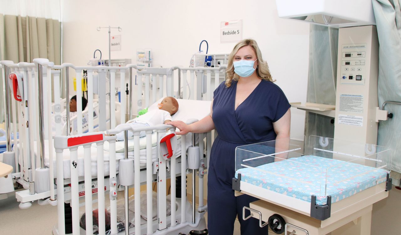 A female Nursing student dressed in blue scrubs stands in a pediatric simulation lab with child patient simulators and medical equipment.