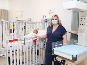 A female Nursing student dressed in blue scrubs stands in a pediatric simulation lab with child patient simulators and medical equipment.