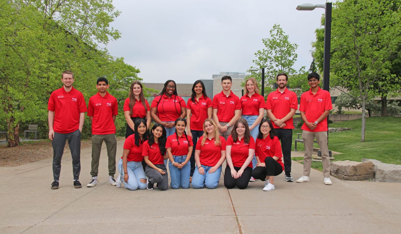 A group of students wearing red shirts stands on an outdoor pathway with buildings behind them to the right and left.