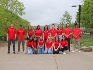A group of students wearing red shirts stands on an outdoor pathway with buildings behind them to the right and left.