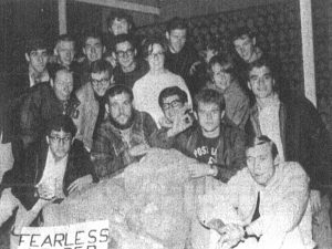 A group of university students gather round a large stone the shape of a military officer’s torso. They hold a sign that reads ‘Fearless leader Sir Isaac.’
