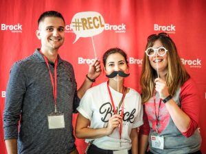 Three people stand in front of a red backdrop that says Brock University on it. The man on the left is holding a #RECL sign, the woman in the centre is holding a fake moustache, and the woman on the right is holding fake glasses.