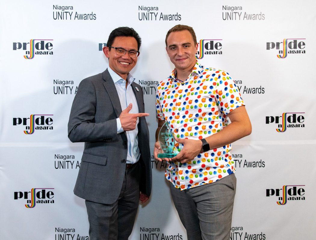 Two men, one holding an award, stand together against a backdrop covered in logos for the Pride Niagara Unity Awards.