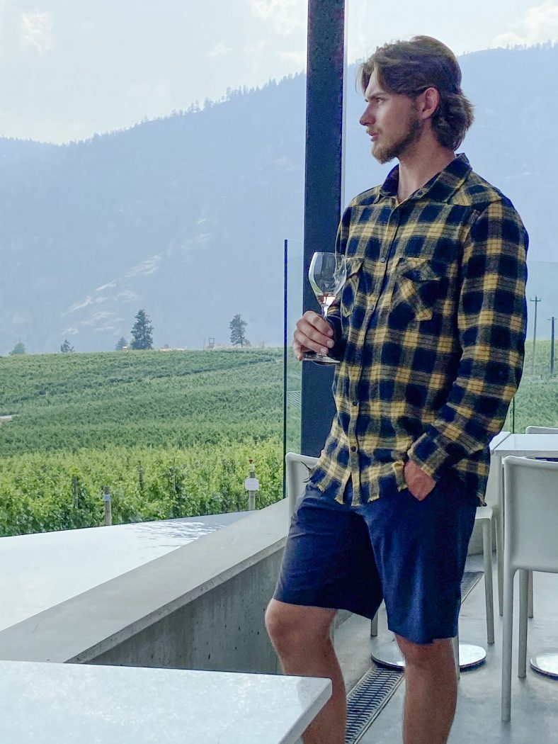 A man poses with a glass of wine in hand inside a winery tasting room with a view of the Rocky Mountains.