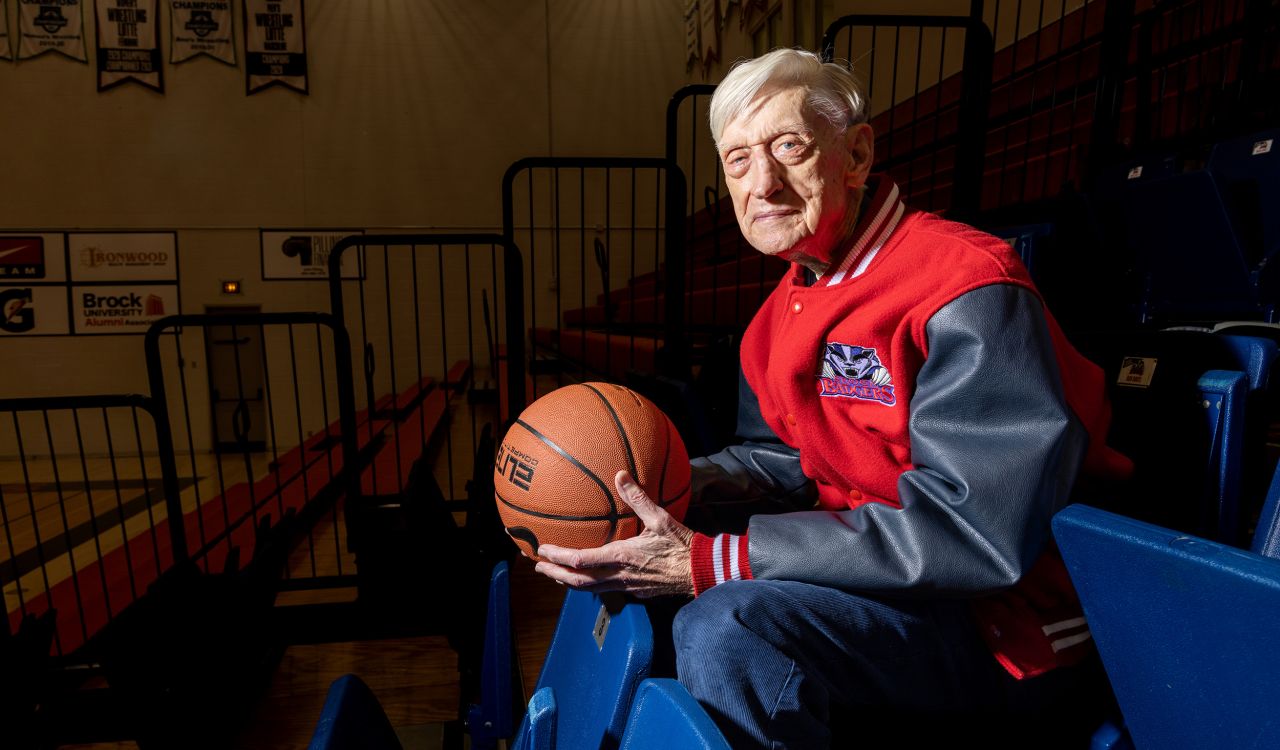 A man in a red letterman's jacket with black sleeves holds a basketball in a darkened gymnasium.