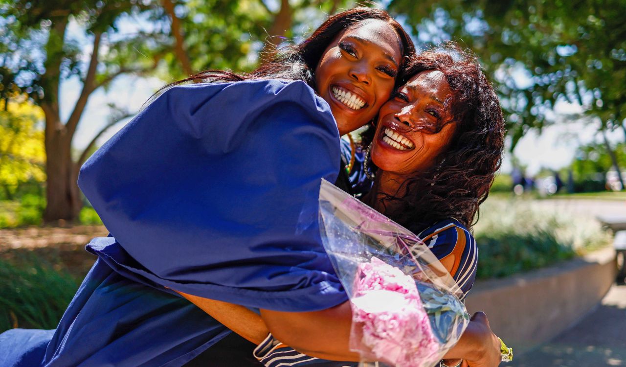 A woman hugs her mom while wearing a blue graduation gown and holding a bouquet of flowers. Both have big smiles on their faces.
