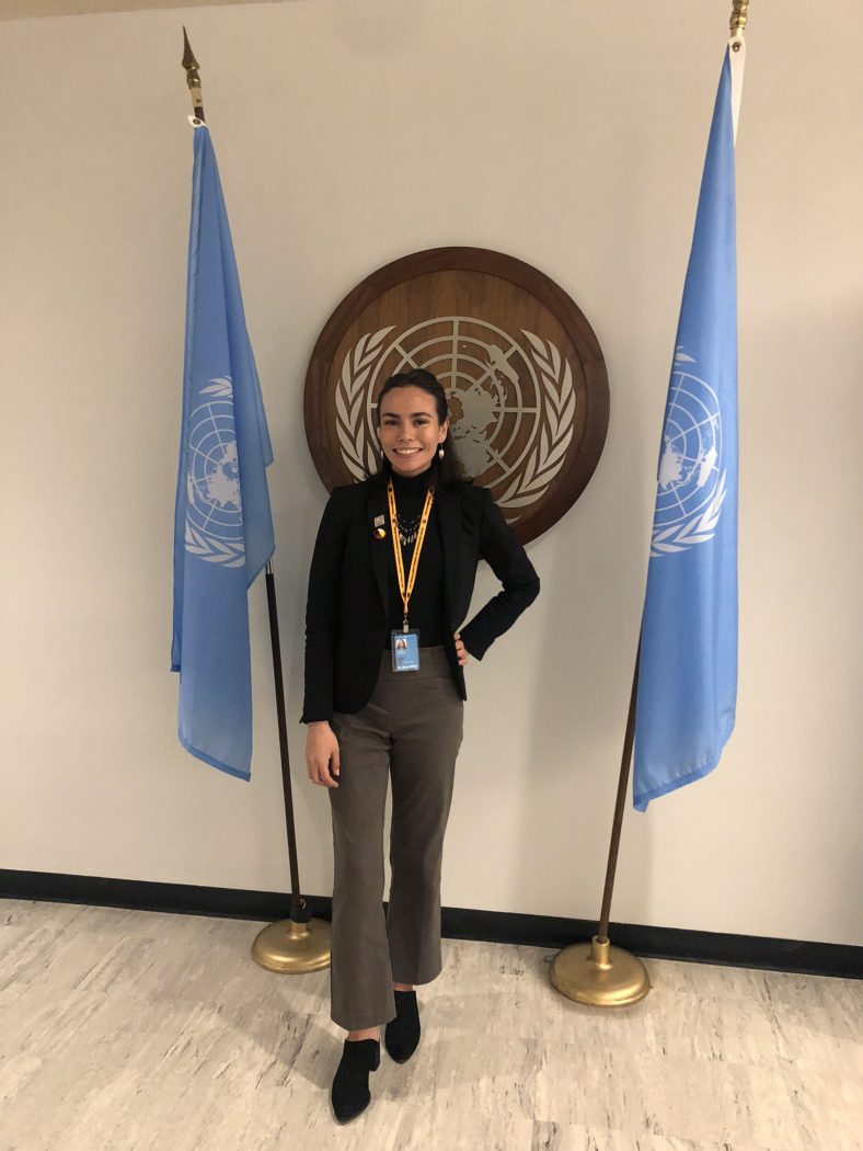 A woman in a black blazer and brown pants stands in front of the seal of the United Nations between two blue United Nations flags.