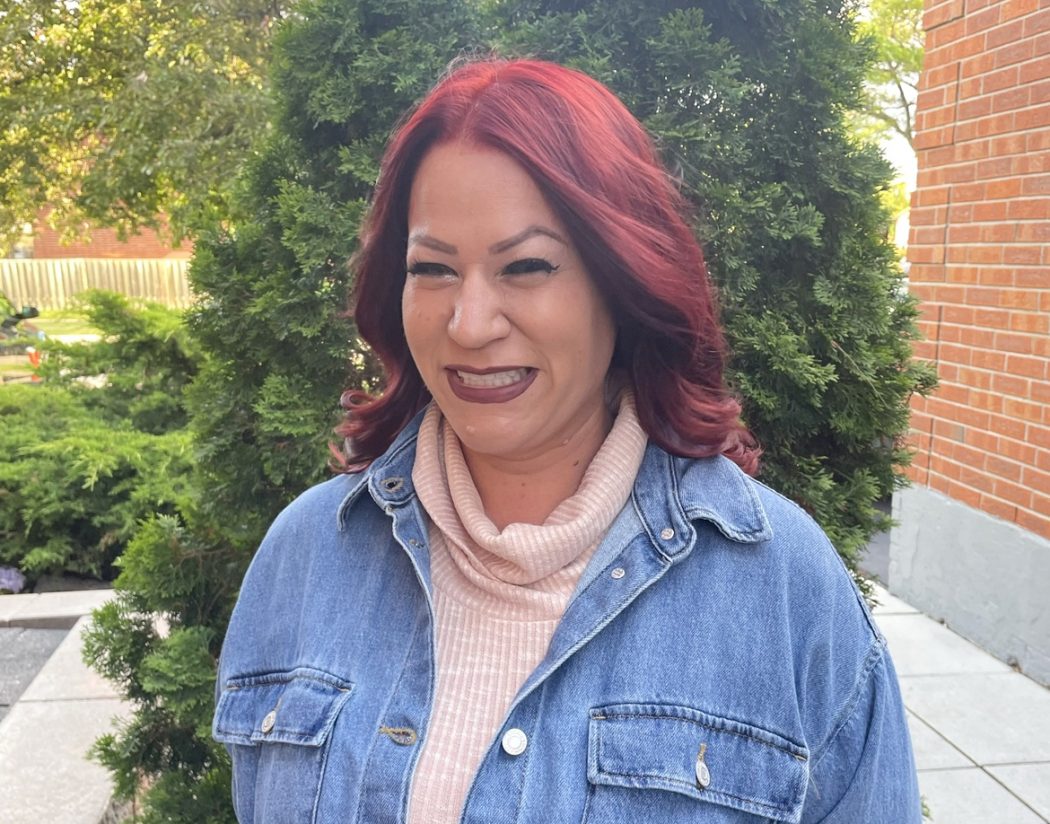 A woman with red hair wearing a jean jacket stands in front of a tree.
