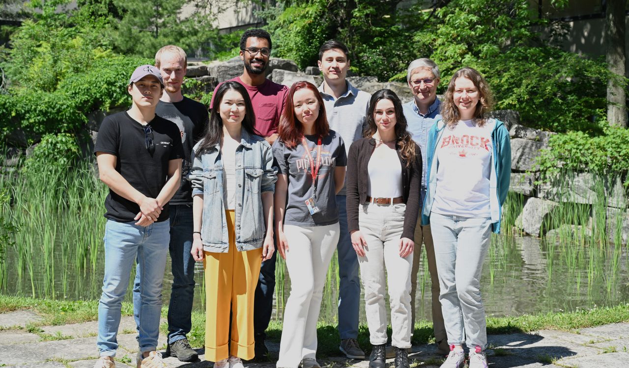 Eight current and former students of Professor Georgii Nikonov stand in two rows in front of Pond Inlet at Brock University. They are surrounded by greenery and backed by a pond and waterfall flowing over grey stone rock wall.