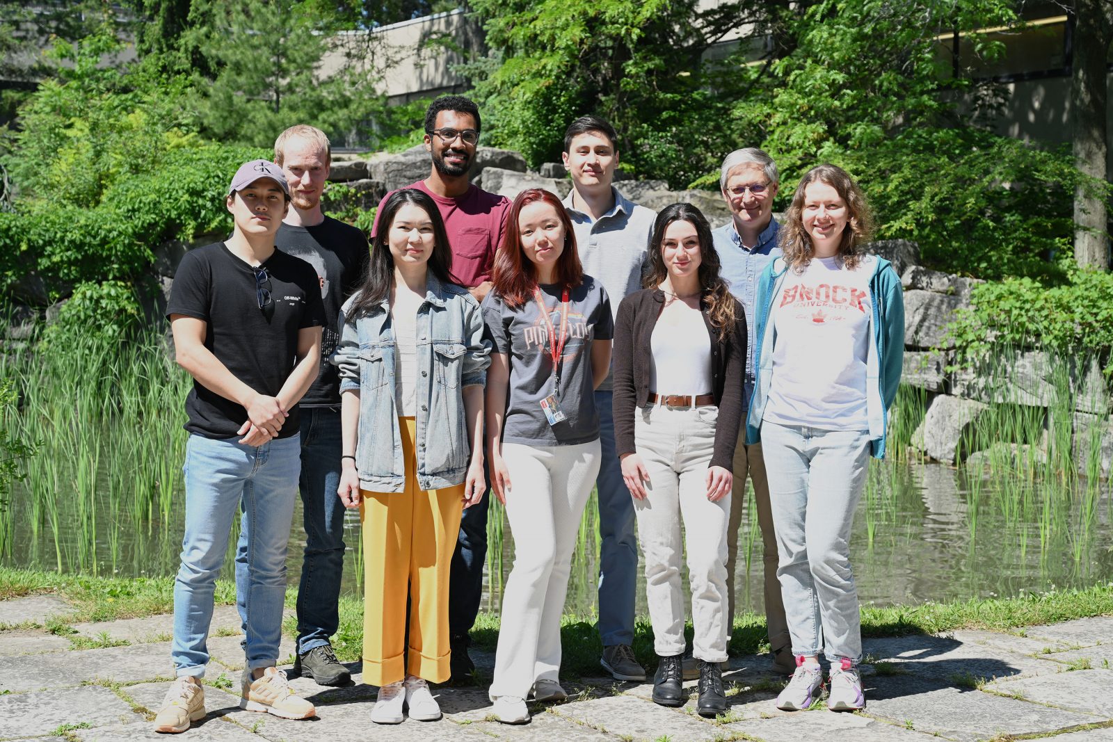 Eight current and former students of Professor Georgii Nikonov stand in two rows in front of Pond Inlet at Brock University. They are surrounded by greenery and backed by a pond and waterfall flowing over grey stone rock wall.
