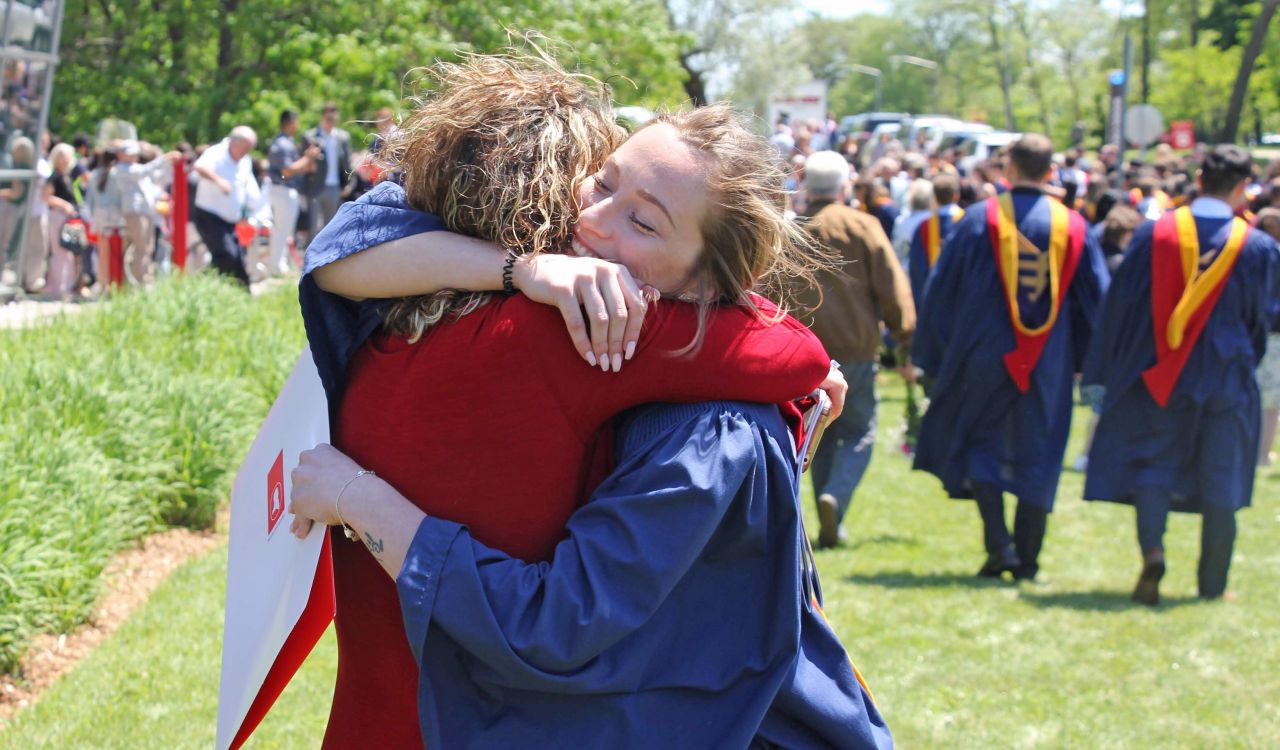 A university graduate in a blue gown hugs a woman while a crowd of graduates walk by in the background.